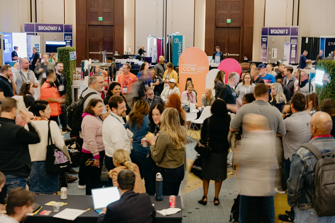 A large group of people gathers and interacts at an indoor conference or trade show, surrounded by booths and informational displays.