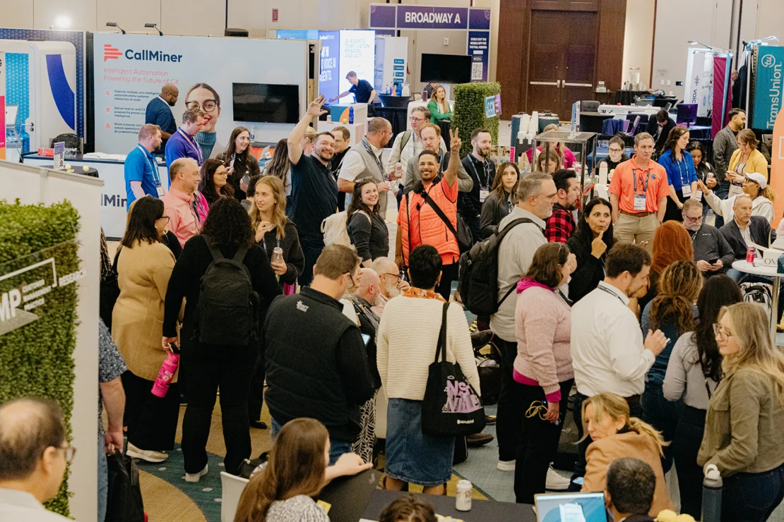 A large group of people gather and interact at a busy conference or trade show, surrounded by booths and displays in a convention hall.