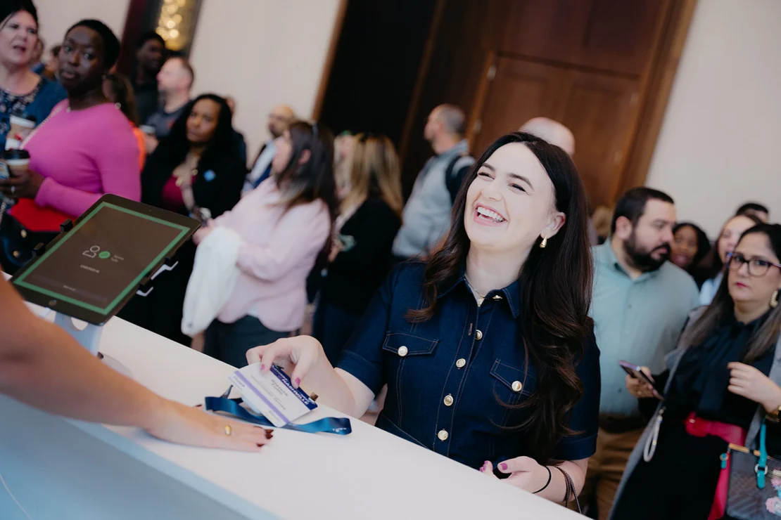 A woman smiles as she checks in at a registration desk while others wait in line behind her at a busy indoor event.