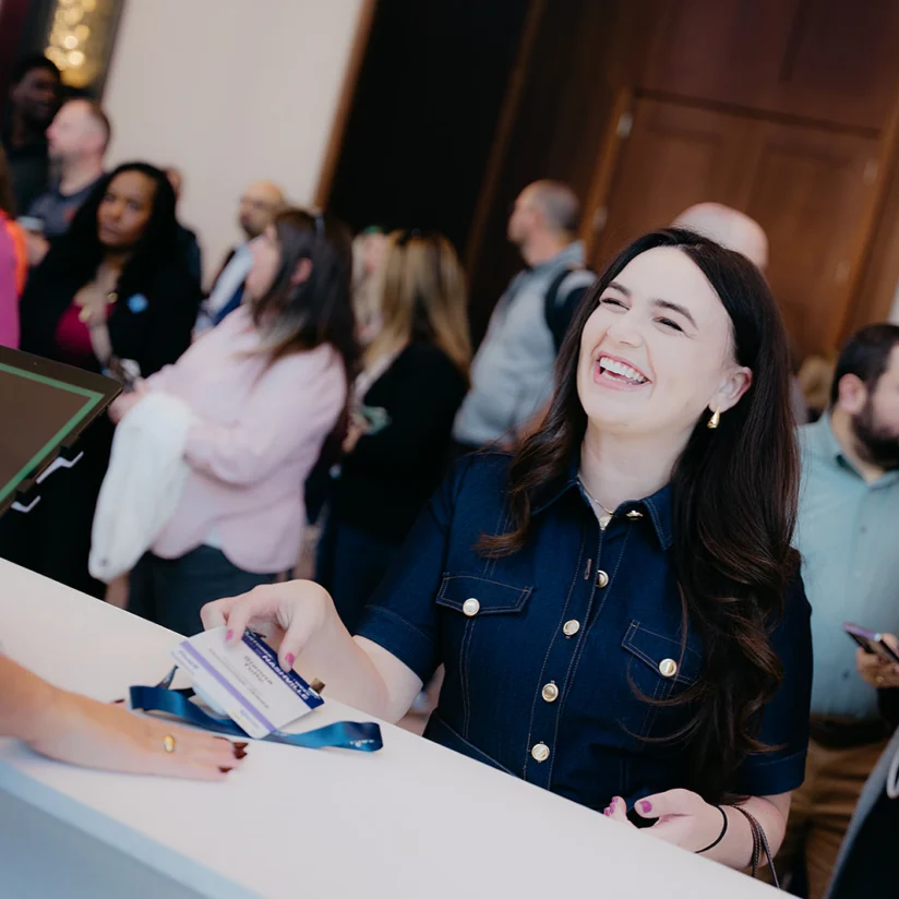 A woman smiles while checking in at a registration desk, holding a badge, with a crowd of people waiting in line behind her.