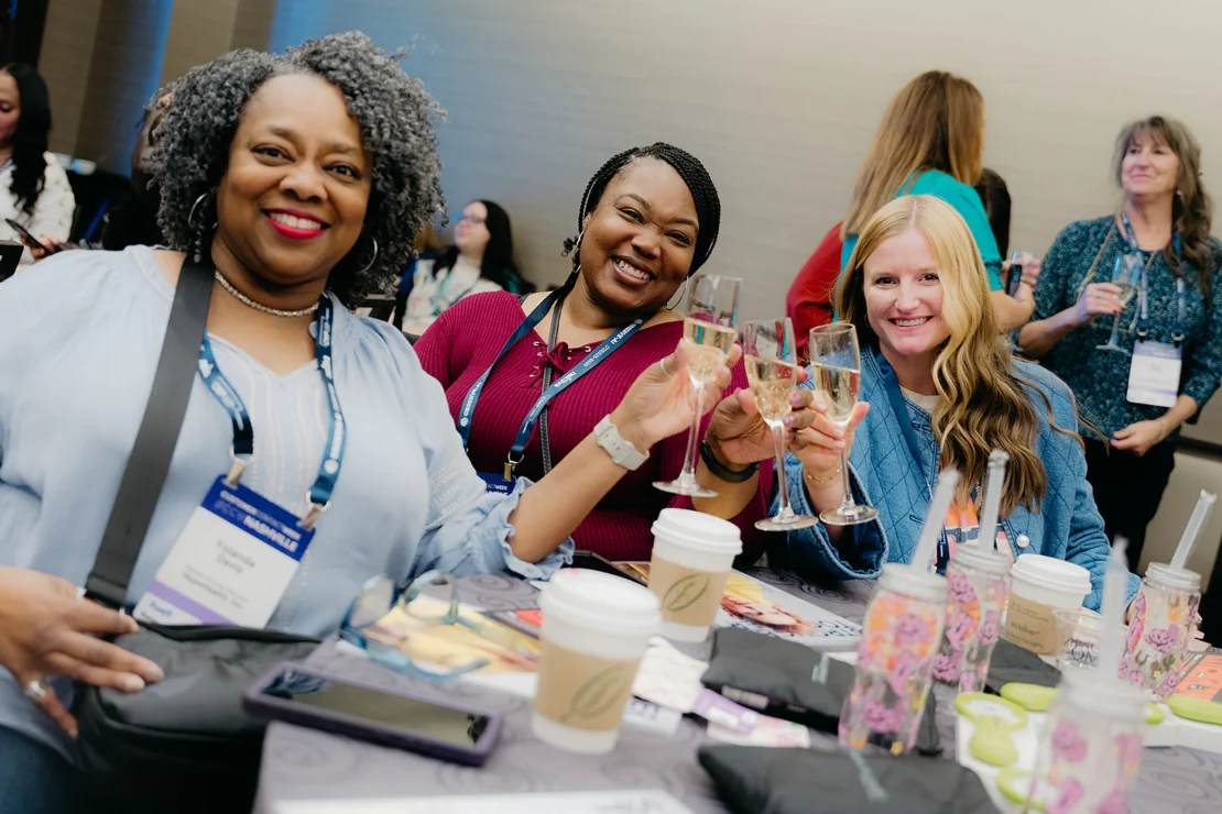 Three women at a conference table smile and toast with glasses of wine, surrounded by coffee cups and name badges, as other attendees mingle in the background.