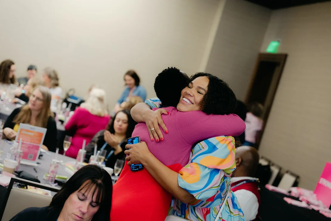 Two women hug and smile in a crowded conference room with several people seated at tables in the background.