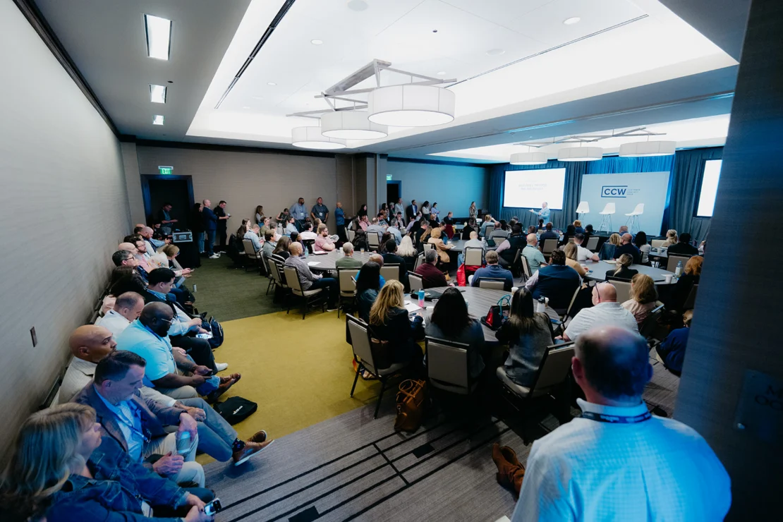 A large group of people seated and standing in a conference room attending a presentation with a speaker at the front near a screen displaying "CCW.