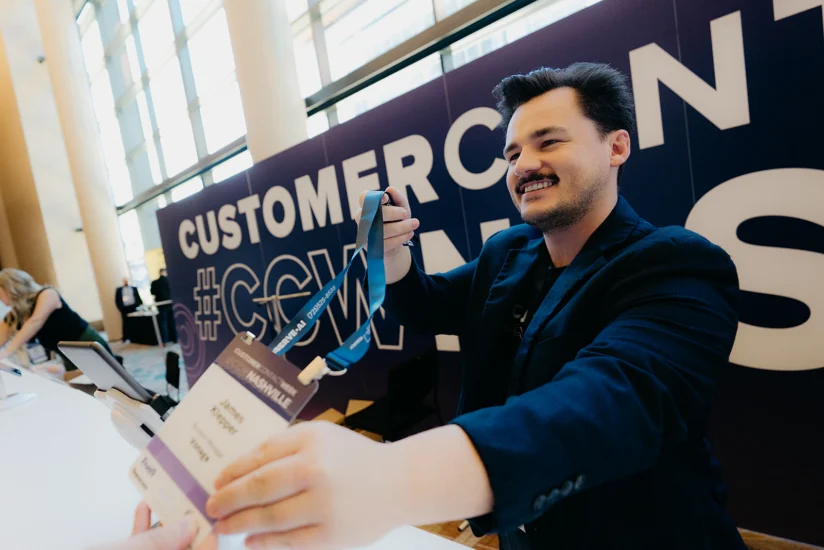 A man hands an attendee a lanyard and name badge at a registration table during a conference. A large "CUSTOMERCON" sign is visible in the background.