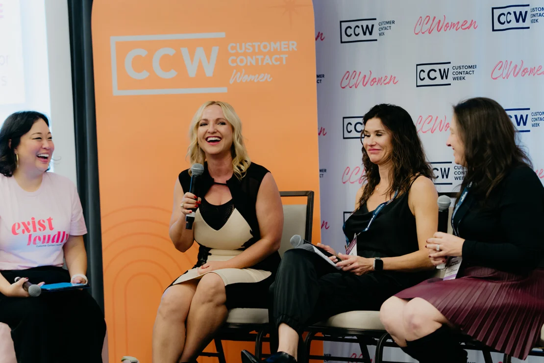 Four women sit and talk on a panel at a Customer Contact Week Women event. One woman holds a microphone and smiles, while the others listen and interact.