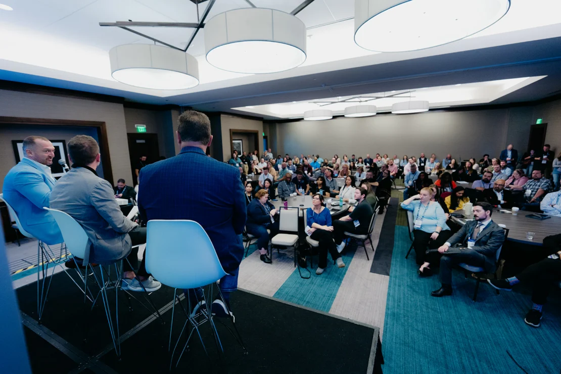 Four people on stage speak to a large seated audience in a conference room during a panel discussion.