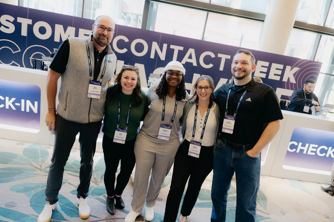 Five people stand side by side smiling at a conference check-in area, all wearing name badges, with a "CHECK-IN" sign visible in the background.