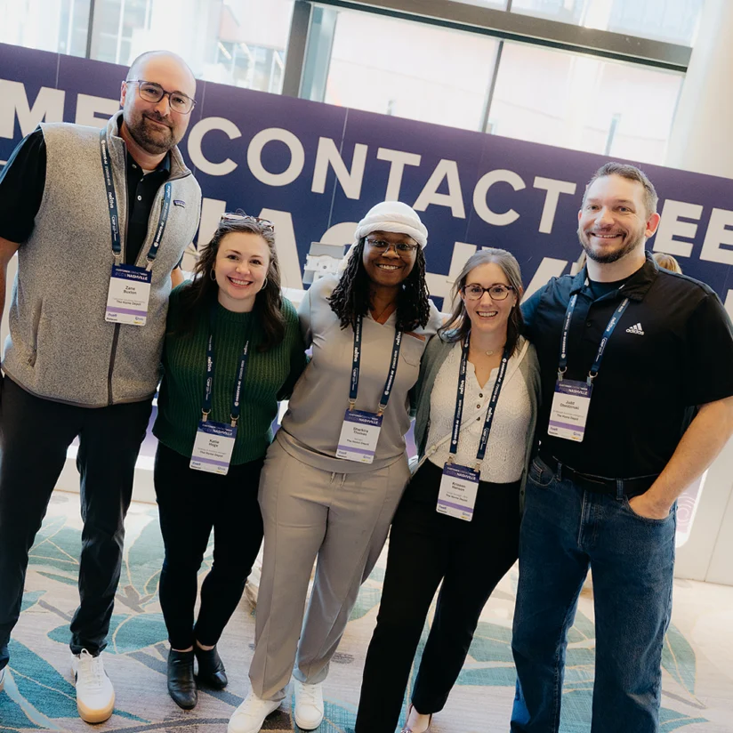 Five people stand side by side smiling at a conference check-in area, all wearing name badges, with a "CHECK-IN" sign visible in the background.