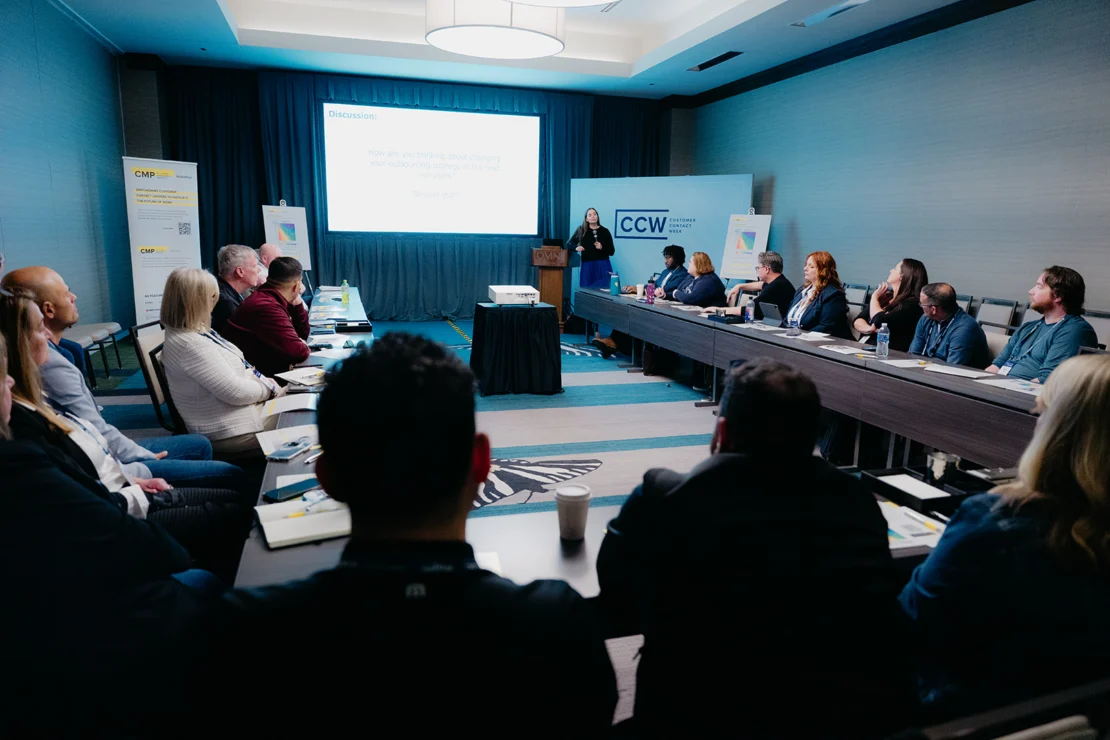 A presenter speaks to a group of people seated around a U-shaped conference table in a meeting room with presentation slides displayed.