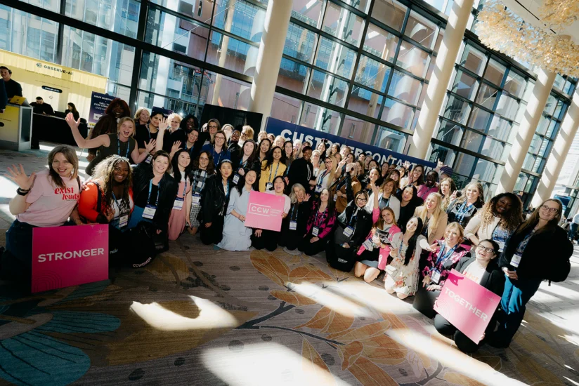 A large group of people, mostly women, pose and smile indoors at an event, holding pink signs that read "STRONGER" and "TOGETHER.