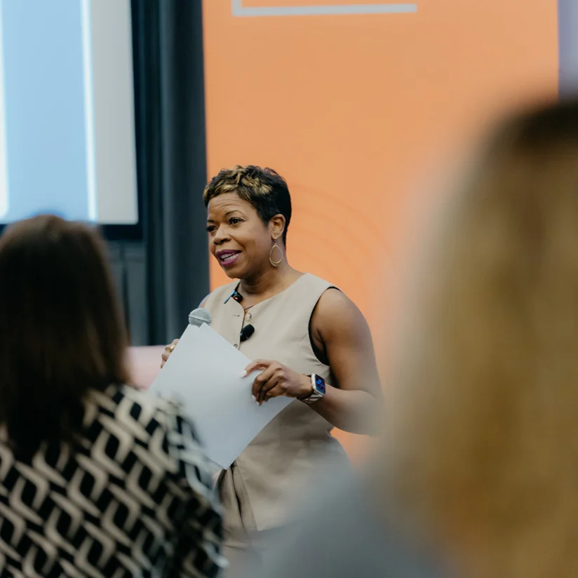 A woman holding papers and a microphone speaks at a Customer Contact Week event, standing in front of an orange banner.