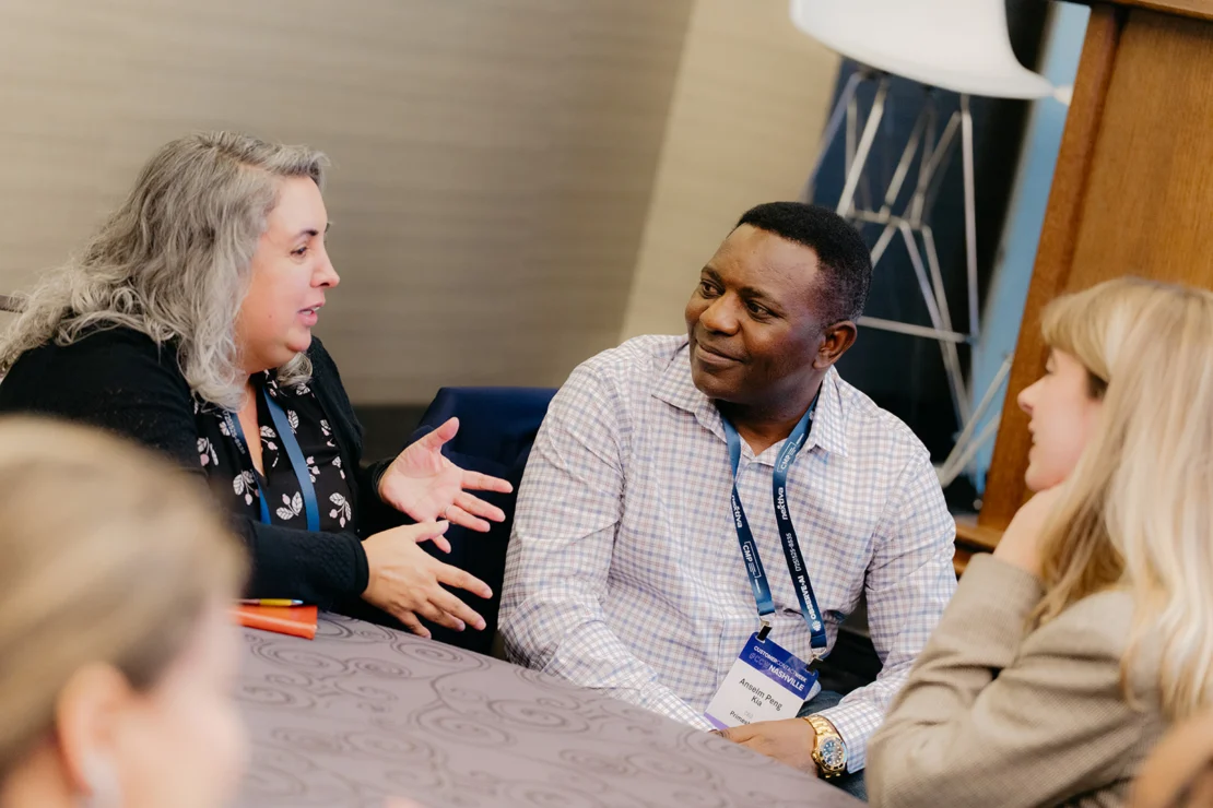 Three people engage in a discussion while seated around a table at a professional event, wearing conference badges.