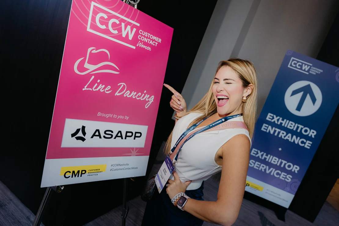 A woman smiles and points at a pink sign for "Line Dancing" at a CCW event, with sponsor logos and an exhibitor entrance sign visible in the background.
