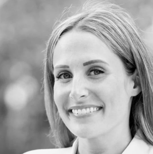 A woman with long, straight hair smiling at the camera, photographed outdoors with a blurred background. The image is in black and white.