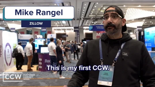 Mike Rangel of Zillow speaks in an exhibit hall at Customer Contact Week, wearing a badge and lanyard, with event booths and attendees in the background.