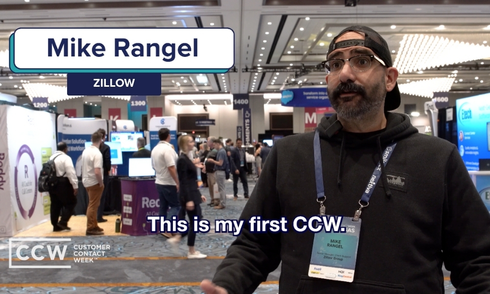 Mike Rangel of Zillow speaks in an exhibit hall at Customer Contact Week, wearing a badge and lanyard, with event booths and attendees in the background.