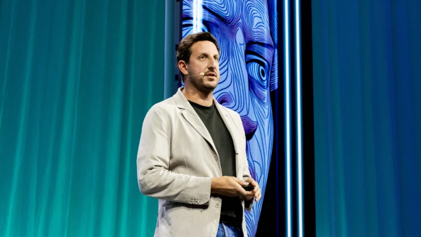 A man with a headset microphone speaks on stage in front of a blue-green curtain and a large screen displaying abstract artwork.