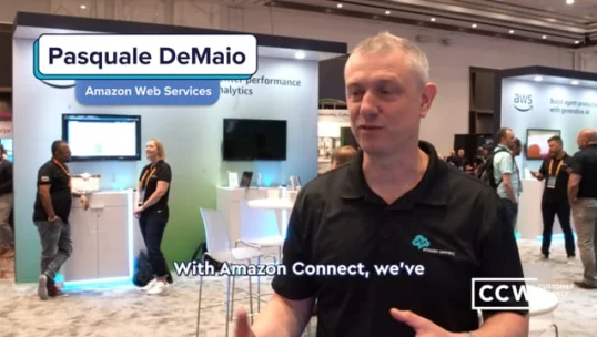 Pasquale DeMaio of Amazon Web Services speaks at a technology conference booth, with other attendees and display screens visible in the background.