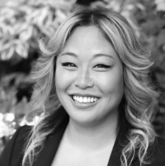 A woman with wavy hair smiles at the camera, wearing a blazer, with foliage and flowers blurred in the background.