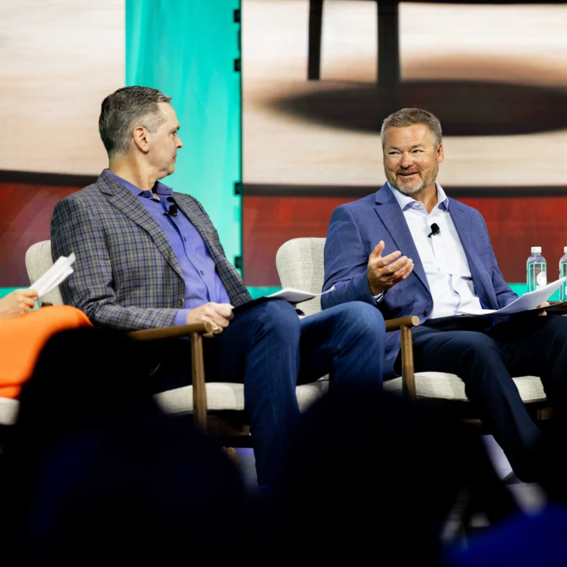 Two men in suits sit on stage chairs during a panel discussion, engaged in conversation, with papers and water bottles in hand.