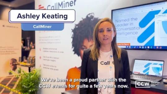 Ashley Keating from CallMiner stands in a conference booth with a screen and banner displaying the company’s branding and achievements.