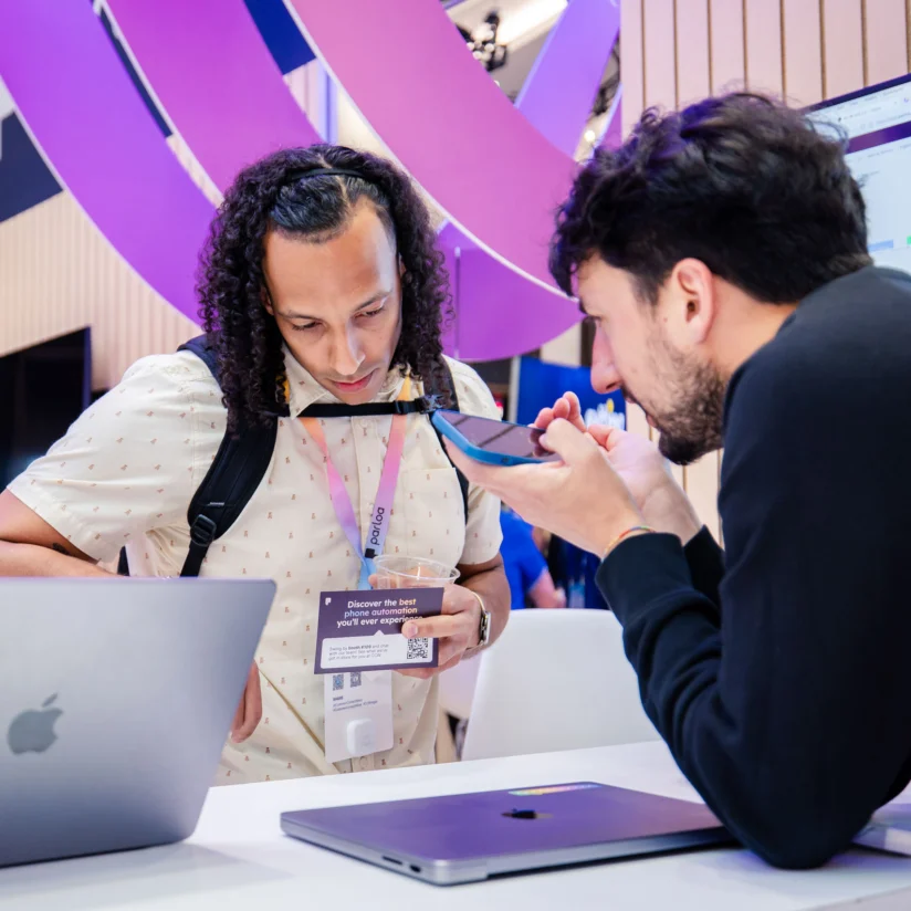 Two men at a tech booth; one is writing on a notepad while the other speaks into his smartphone. Laptops and a monitor are visible on the table.