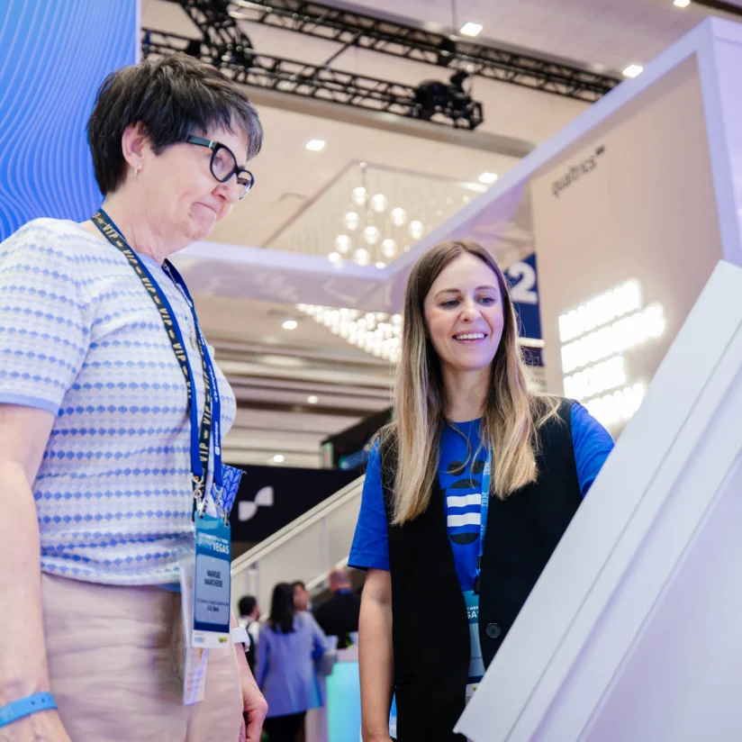 Two women stand at a digital kiosk in a convention hall, one interacting with the screen while the other looks on. Trade show booths and signage are visible in the background.