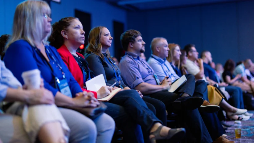 A group of people sit in rows, attentively listening to a presentation at a conference or seminar in a dimly lit room.
