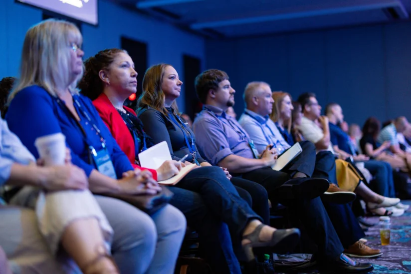 A group of people sit in rows, attentively listening to a presentation at a conference or seminar in a dimly lit room.