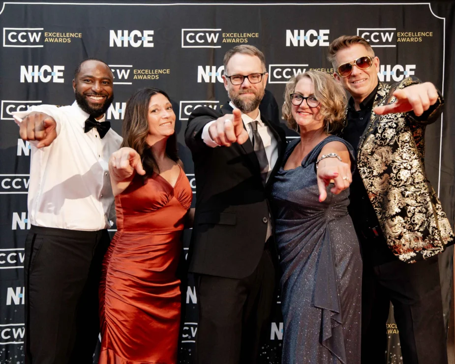 Five people dressed formally stand in front of a CCW Excellence Awards and NICE backdrop, smiling and pointing toward the camera.