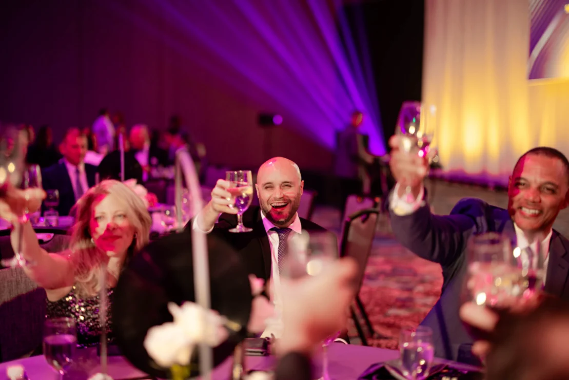 A group of people in formal attire sit at a table, raising glasses in a toast at a banquet or gala with colorful lighting in the background.