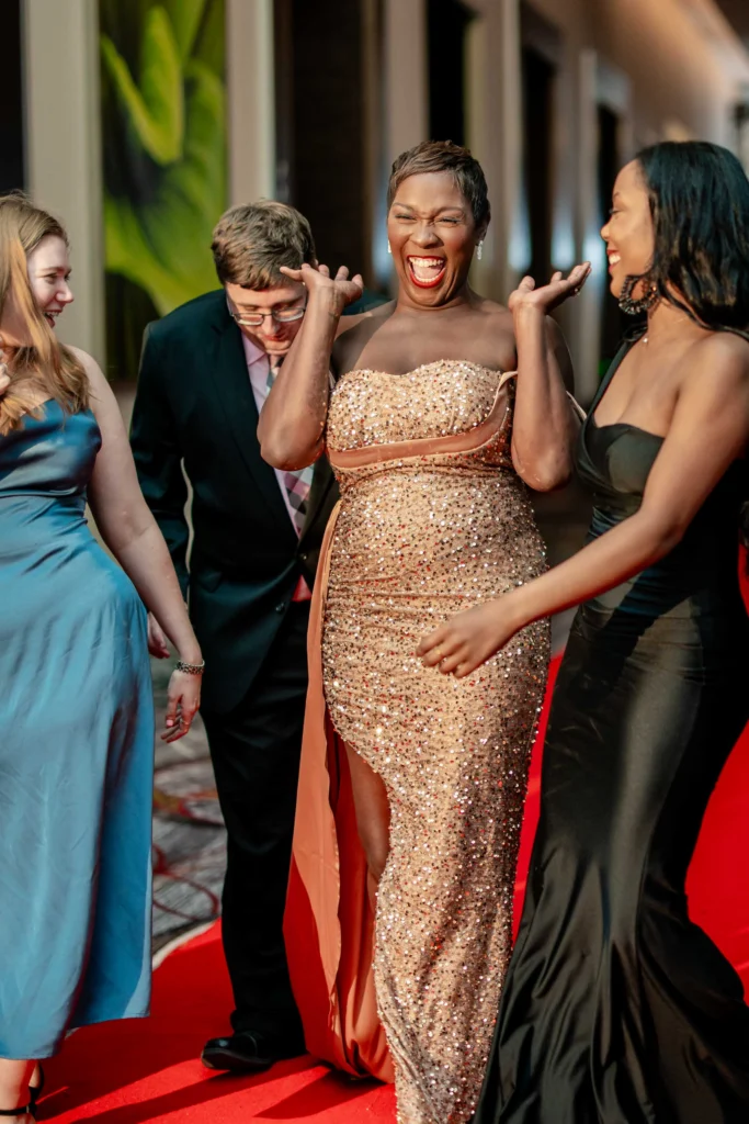 Four people dressed in formal attire stand on a red carpet. The woman in the center, wearing a sequined gown, smiles and gestures with excitement.
