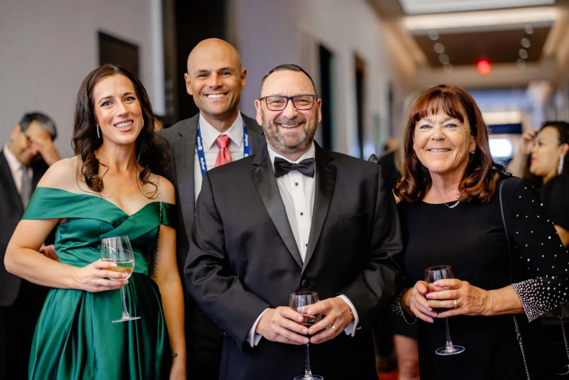 Four adults dressed in formal attire pose together at an indoor event, each holding a glass of wine and smiling at the camera.