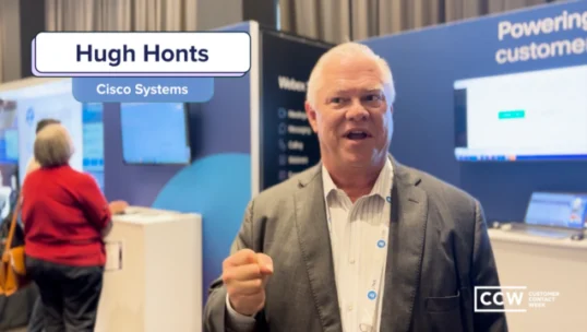 A man in a suit stands in a conference booth area, holding his fist up. A name tag on the image reads “Hugh Honts, Cisco Systems.” CCW Customer Contact Week branding is visible.