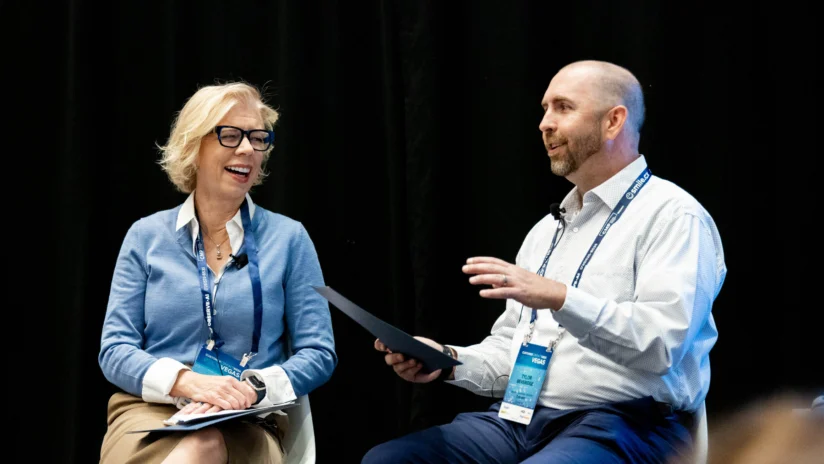 Two people sit on stage conversing, both wearing conference badges and microphones, with one holding a folder and the other smiling.