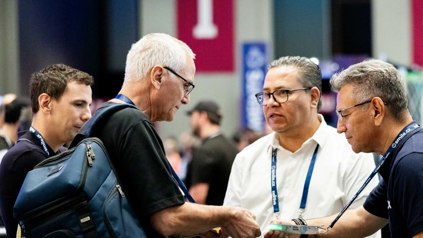 Four men stand together at a conference, engaged in conversation; one holds a book and another carries a blue backpack.