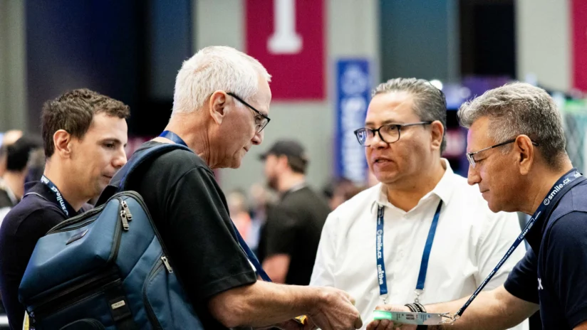 Four men stand together at a conference, engaged in conversation; one holds a book and another carries a blue backpack.