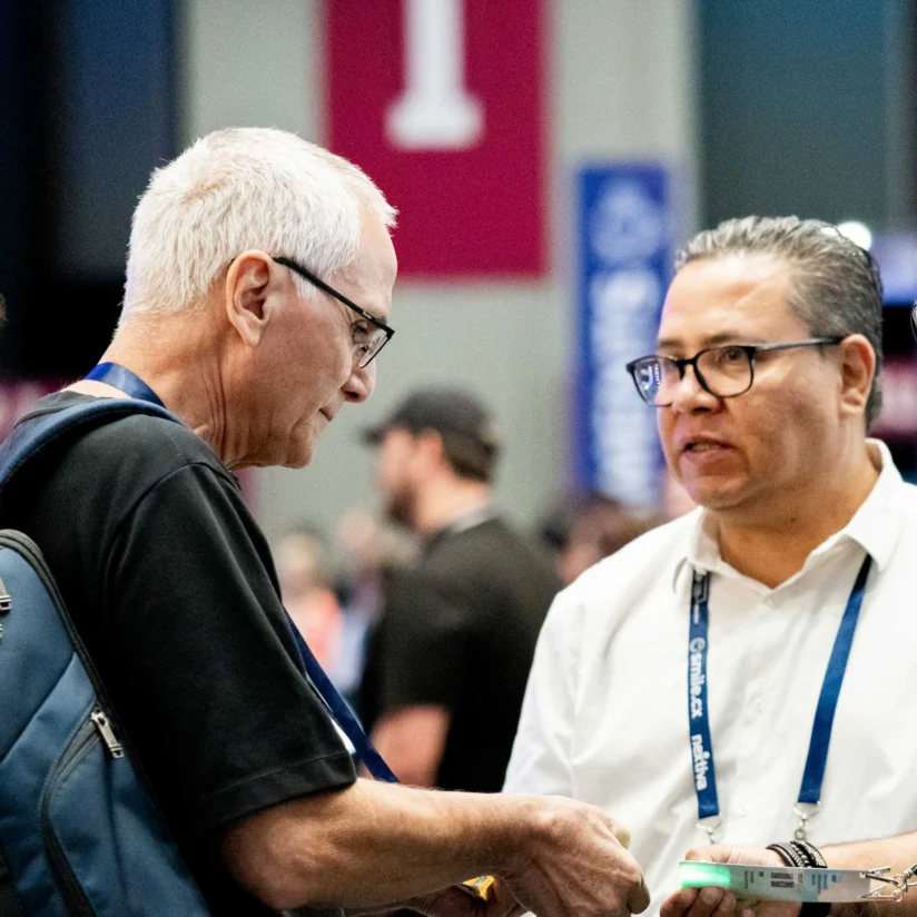 Four men stand together at a conference, engaged in conversation; one holds a book and another carries a blue backpack.