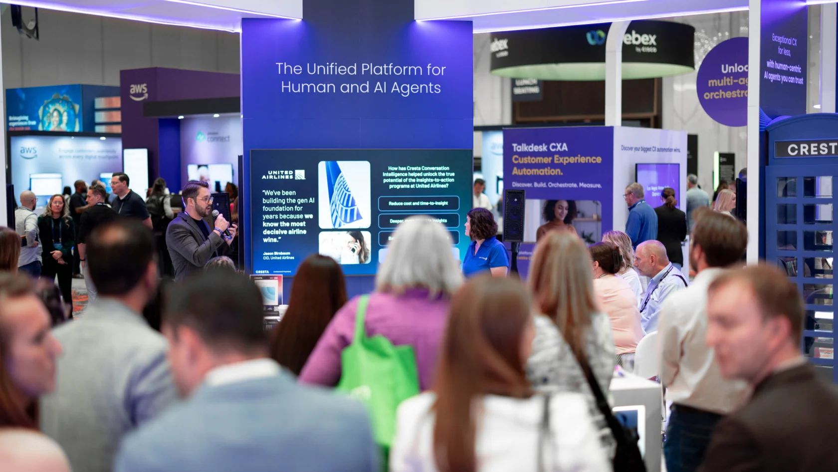 A group of people attend a tech conference, gathered around a booth with a screen displaying information about AI and human agent platforms.