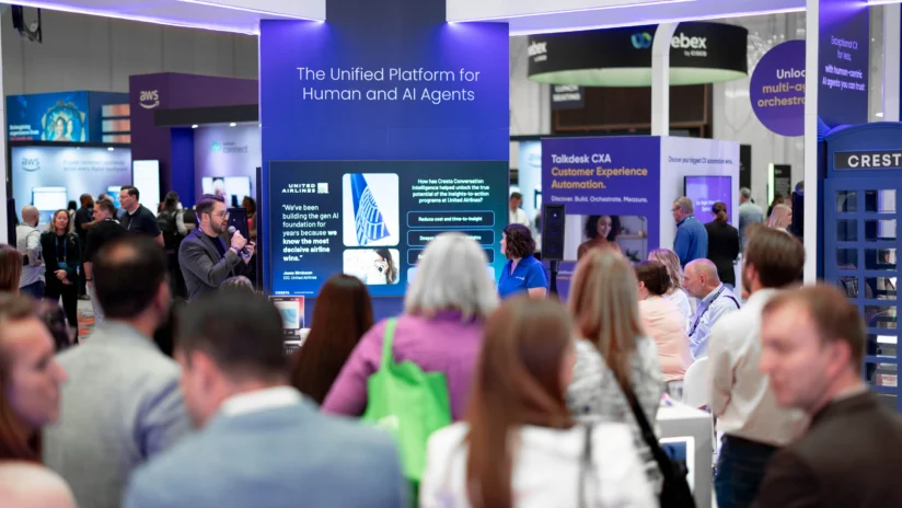 A group of people attend a tech conference, gathered around a booth with a screen displaying information about AI and human agent platforms.