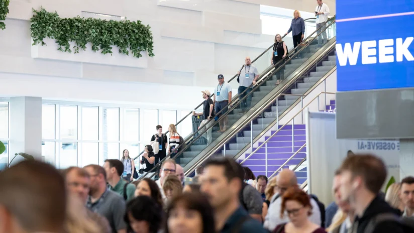 A crowd of people gathers indoors while others ride an escalator in a brightly lit convention center.