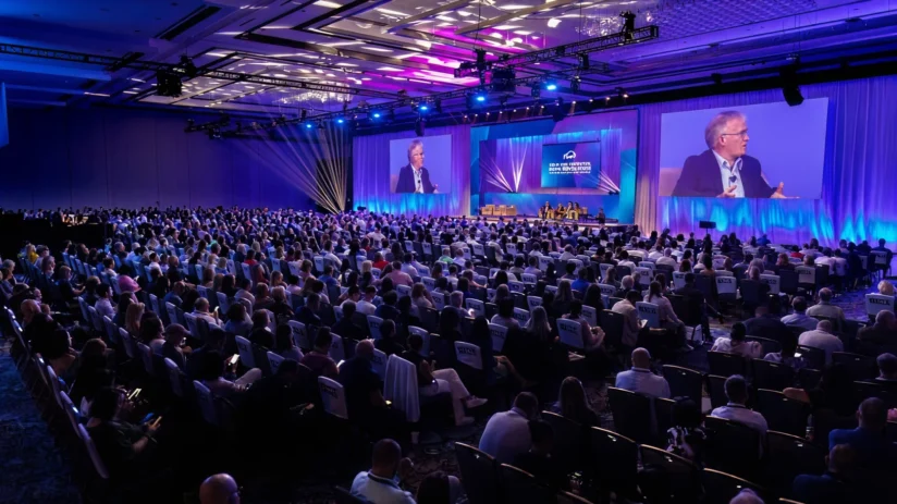Large audience seated in a conference hall watching a panel discussion on stage; presenter speaking, event lighting in blue and purple tones, with presentation projected on big screens.