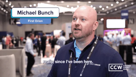 A bald man wearing a navy polo shirt with "First Orion" branding stands in a conference hall, speaking in an interview. Text overlay shows "Michael Bunch, First Orion" and "CCW Customer Contact Week.