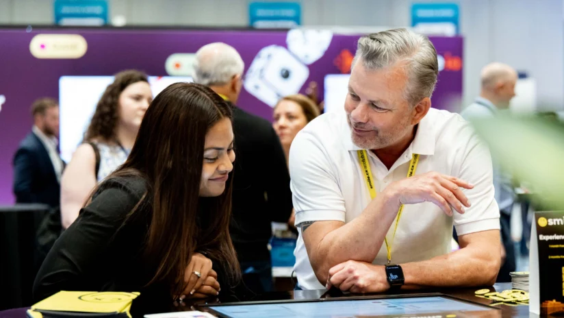 A man and a woman look at a tablet together at an event booth, with other people and informational displays visible in the background.