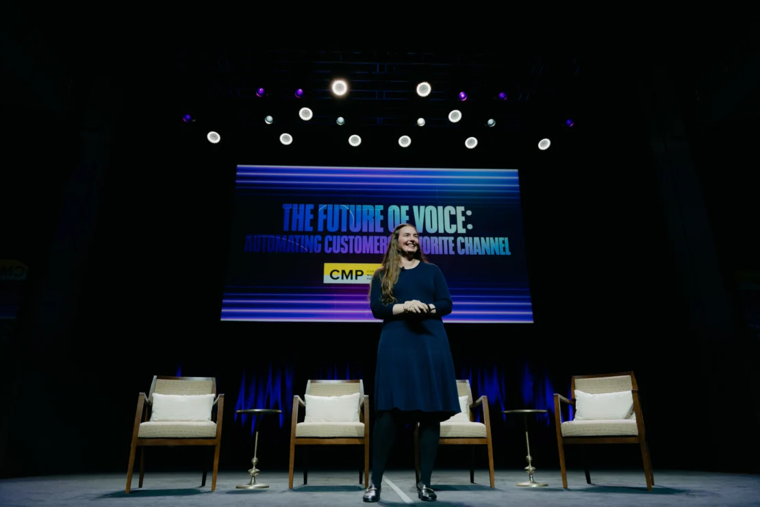 A woman stands on stage in front of a screen displaying "The Future of Voice: Automating Customers' Favorite Channel" with four empty chairs beside her.