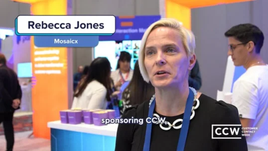 A woman with short blonde hair speaks at a conference booth, wearing a blue lanyard and black necklace. Other attendees and event signage are visible in the background.