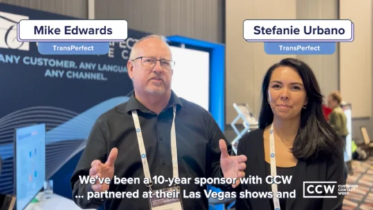 Two people wearing conference badges stand indoors, speaking to the camera in front of a display. Text on screen identifies them as Mike Edwards and Stefanie Urbano from TransPerfect.