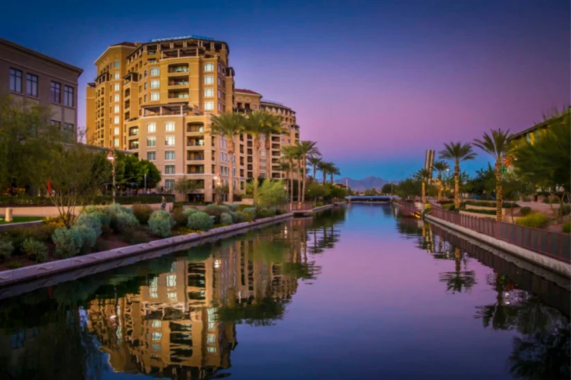 Canal lined with palm trees at sunset in Scottsdale, Arizona, with a residential building reflected in the water and mountains visible in the distance under a purple and blue evening sky.
