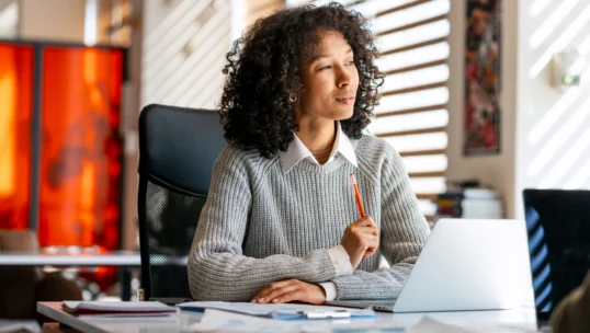 Black woman professionally dressed, sitting at desk, holding a red pencil, looking away and thinking in a sunny office