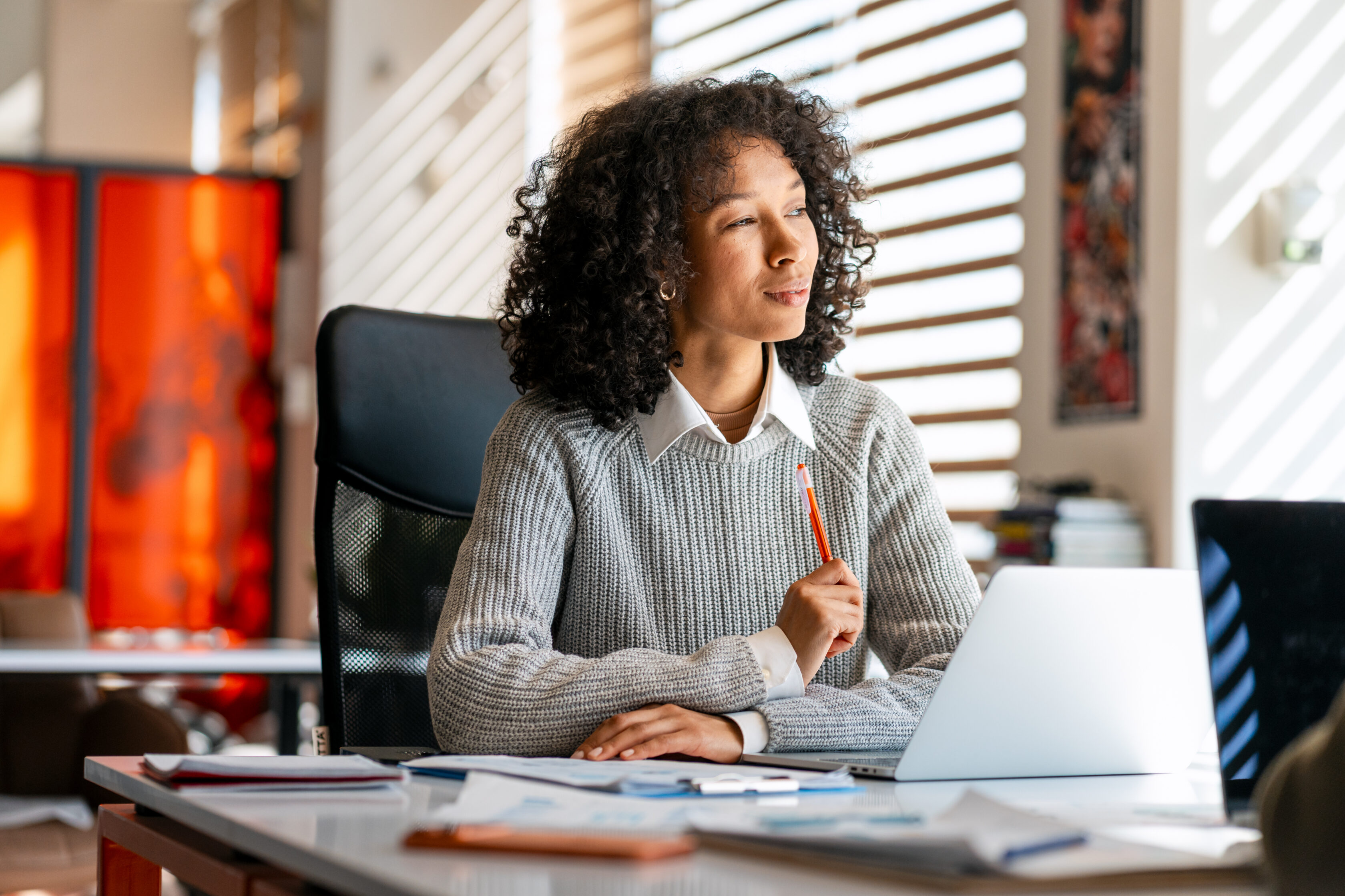 Black woman professionally dressed, sitting at desk, holding a red pencil, looking away and thinking in a sunny office